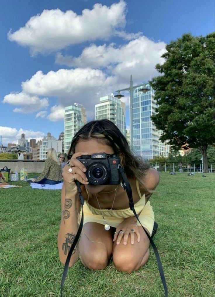 a woman taking a selfie with her camera in the grass near some tall buildings