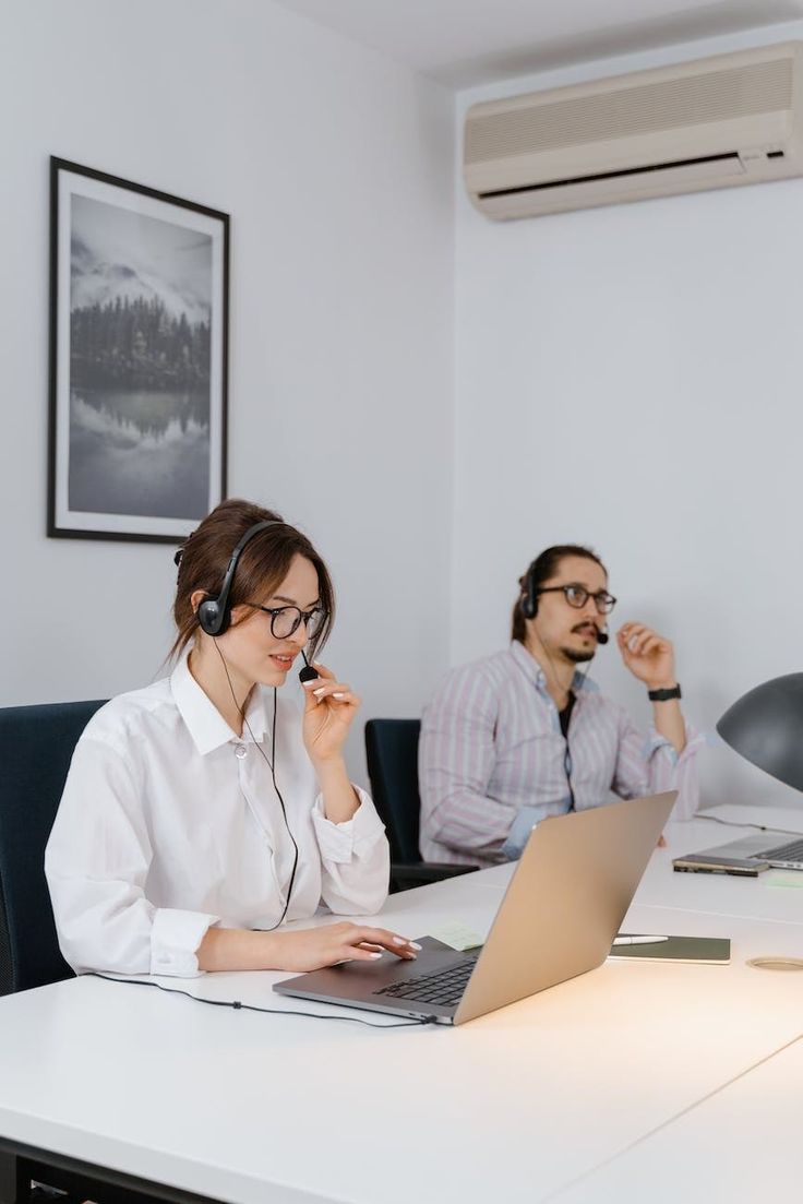 two people sitting at a table with laptops and headphones on, one is talking on the phone