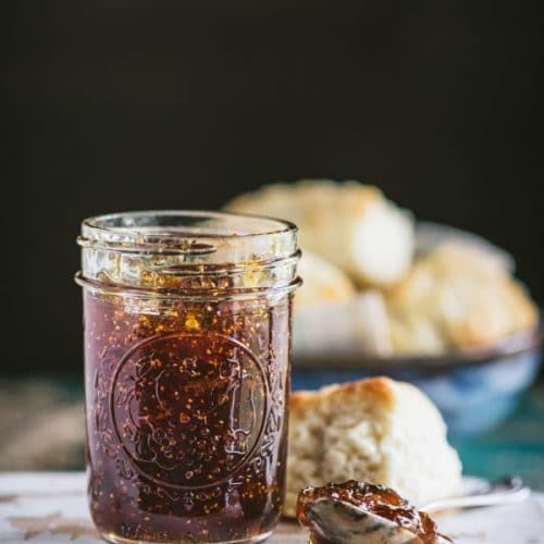 a glass jar filled with jam sitting on top of a wooden table next to muffins