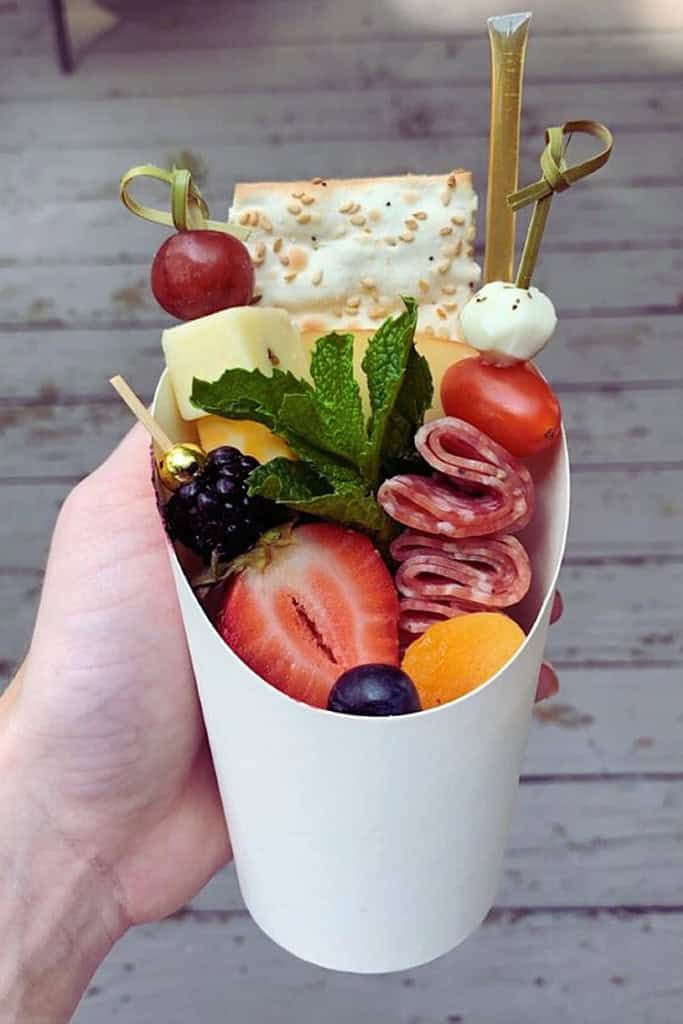 a person holding up a cup filled with fruit and cheese on top of a wooden table