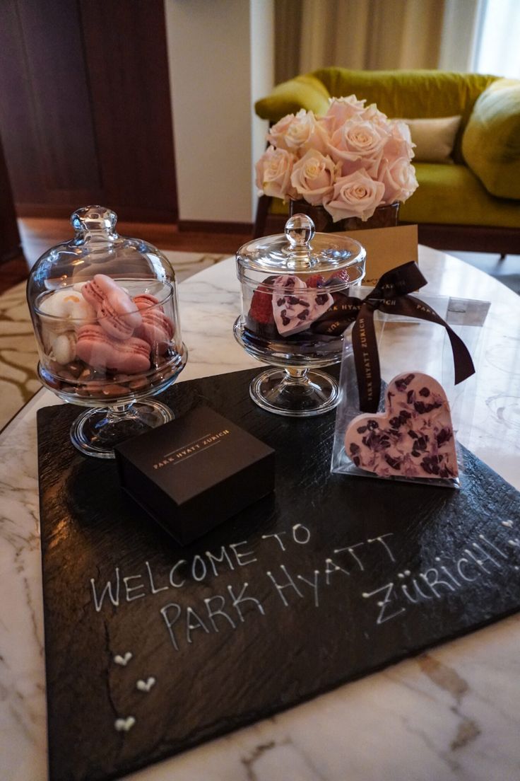 a welcome sign is on the table with some candies in glass containers and flowers