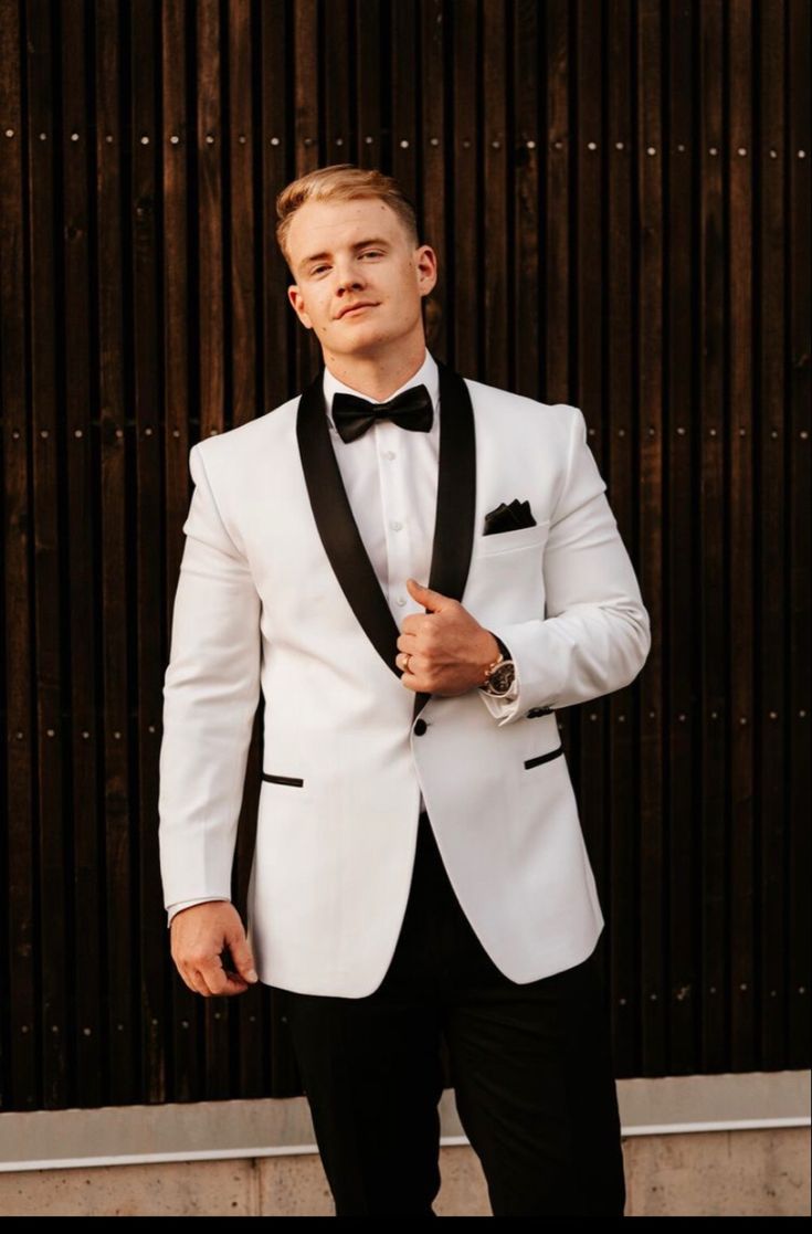 a man in a white tuxedo and black bow tie standing next to a wall