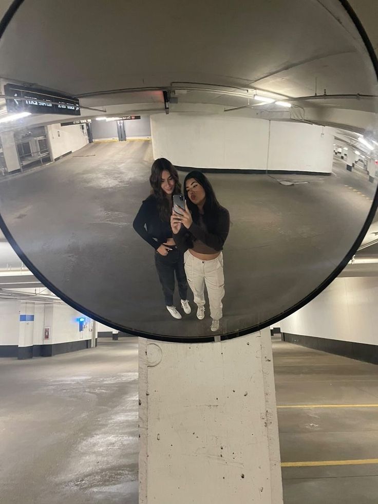 two women taking a selfie in front of a round mirror at an indoor parking garage