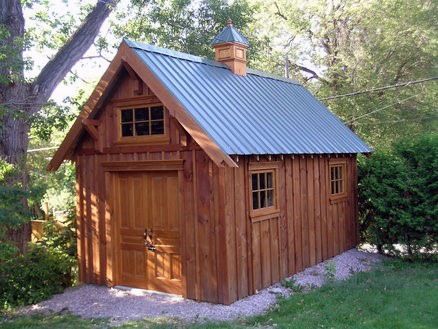 a small wooden shed with a metal roof and windows on the side of it, surrounded by green grass and trees