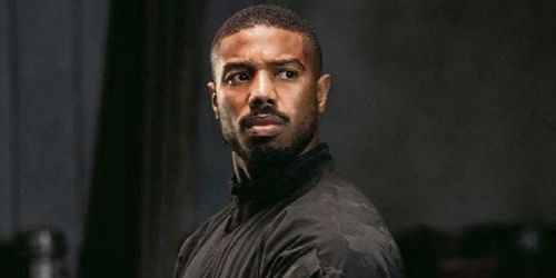 a man with a beard standing in front of a black curtain and looking at the camera