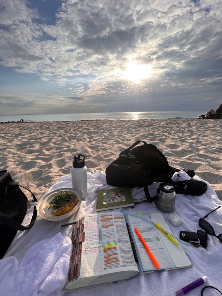 an open book on a beach next to a backpack and some other items in front of the sun