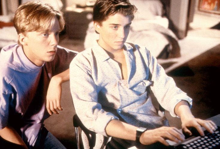 two young men sitting in front of a computer keyboard and monitor looking at each other