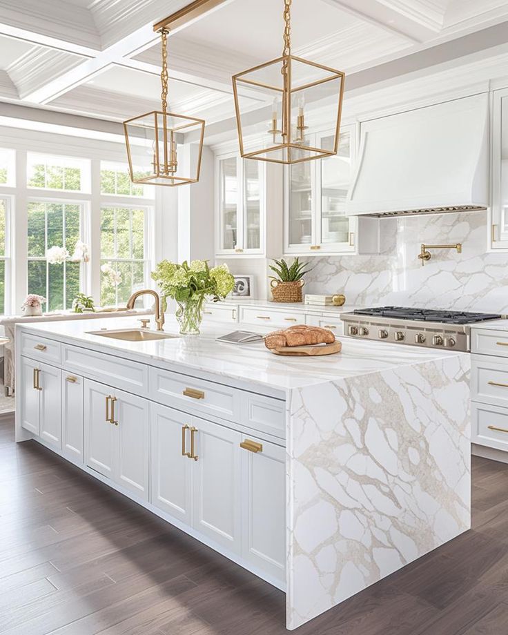 a white kitchen with marble counter tops and gold accents on the island, along with hanging lights