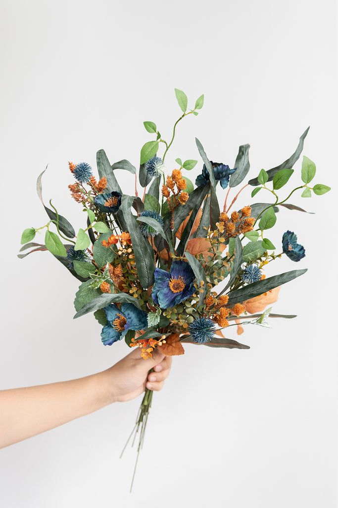 a person holding a bouquet of flowers with green leaves and oranges on it in front of a white background