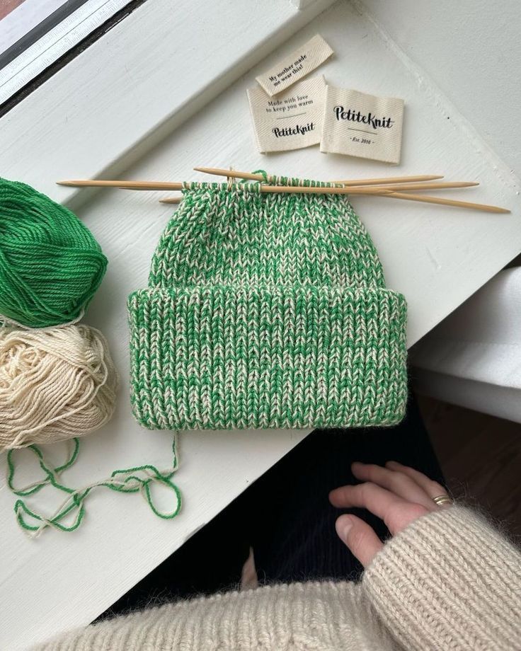 a green knitted beanie sits next to yarn and knitting needles on a white table