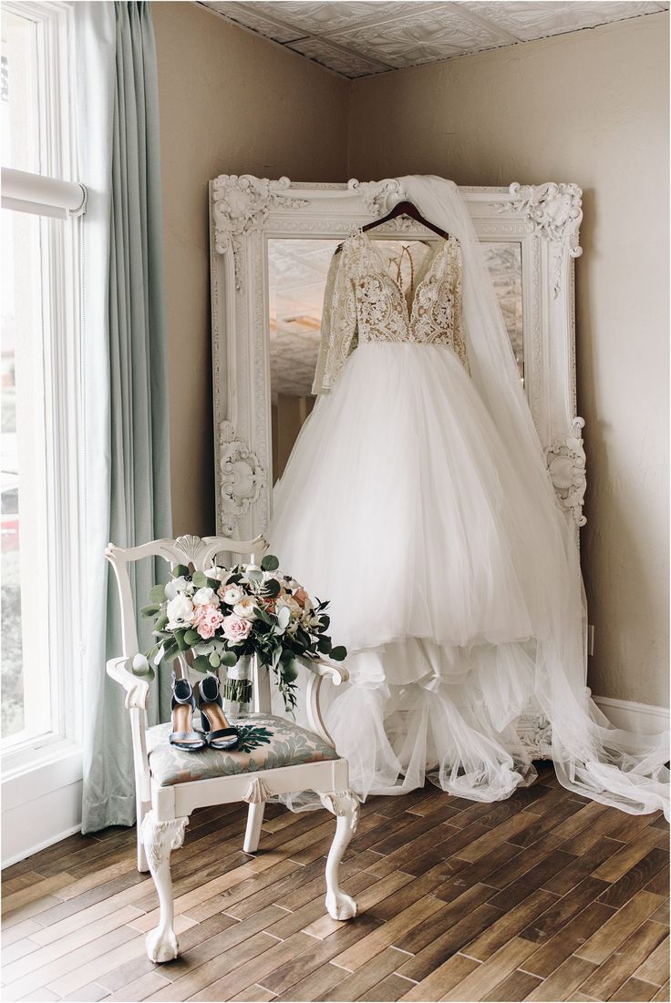 a wedding dress hanging in front of a mirror with flowers on the floor next to it