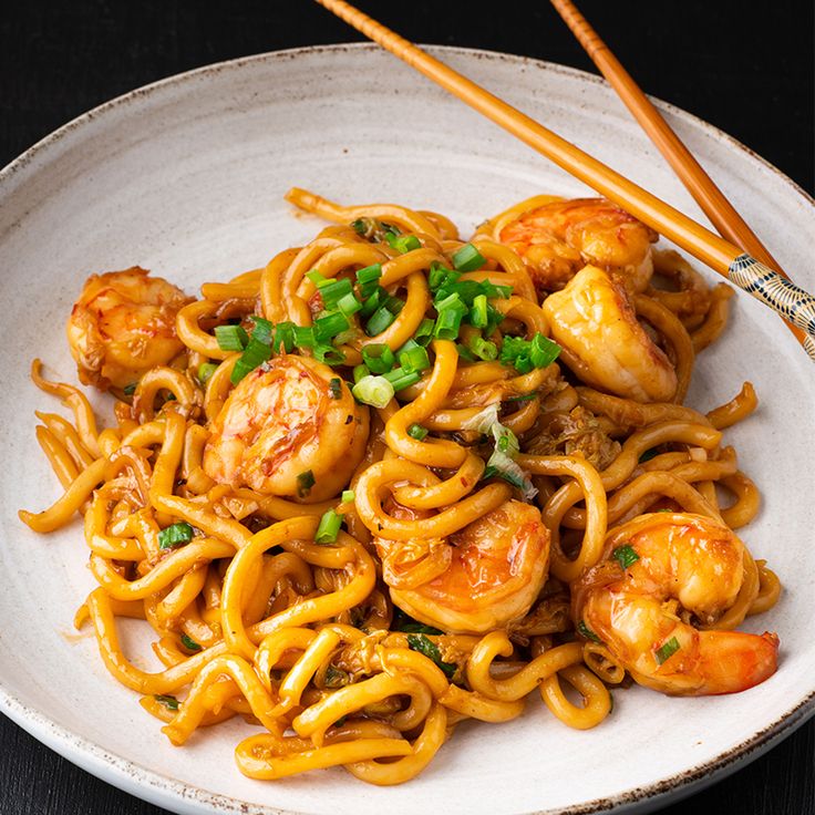 a white plate topped with noodles and shrimp next to chopsticks on a table