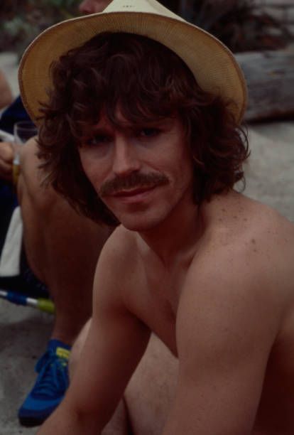 a shirtless man sitting on the beach wearing a hat