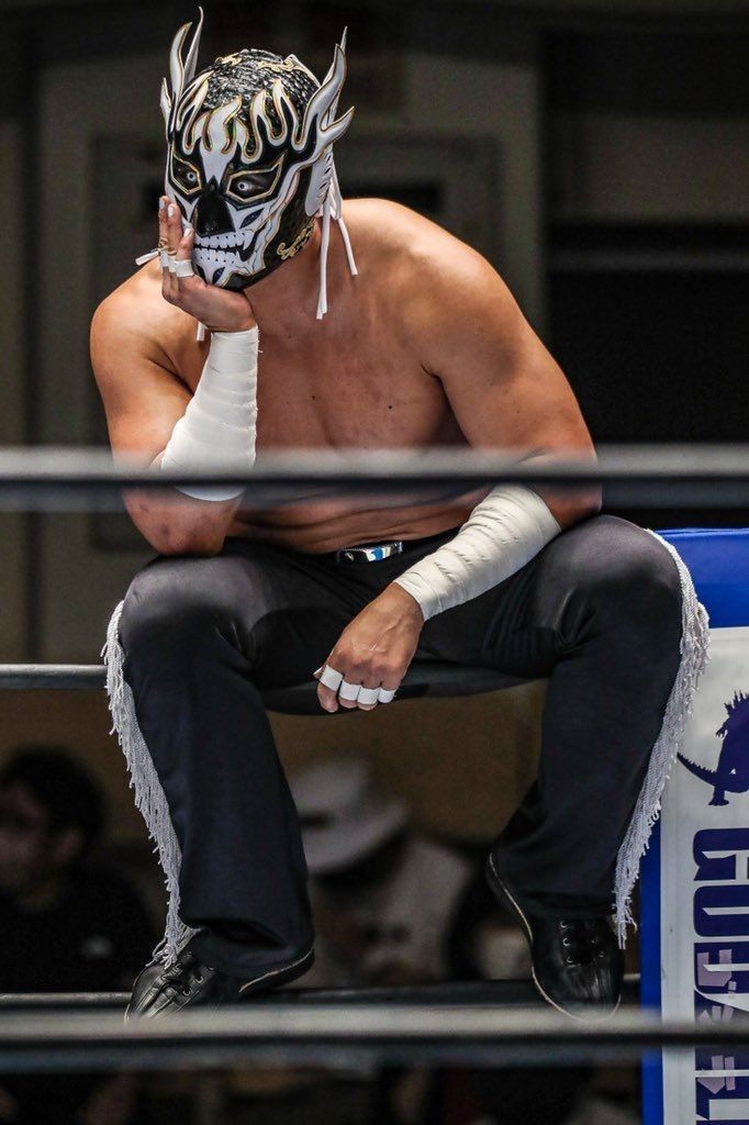 a wrestler sitting on the edge of a wrestling ring wearing a mask and holding his hand to his face