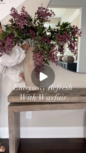 a woman standing next to a wooden table holding a vase with purple flowers on it