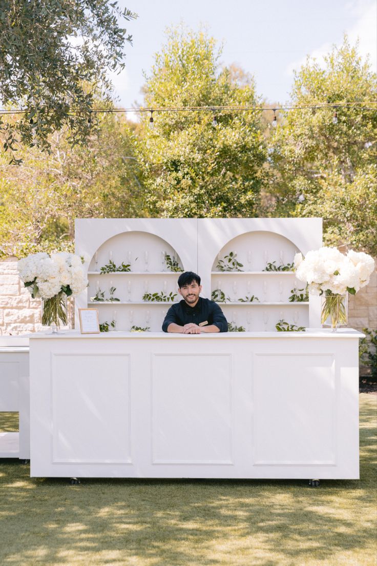 a man standing behind a white counter with flowers on it in front of some trees