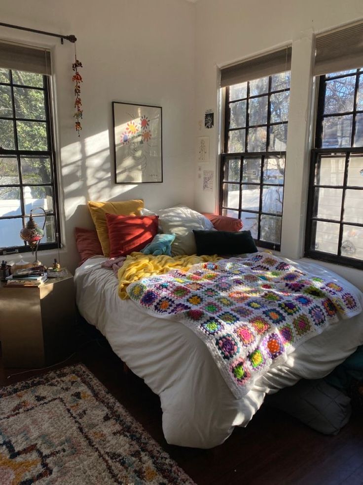 a bedroom with two large windows and a bed in the middle, surrounded by colorful pillows