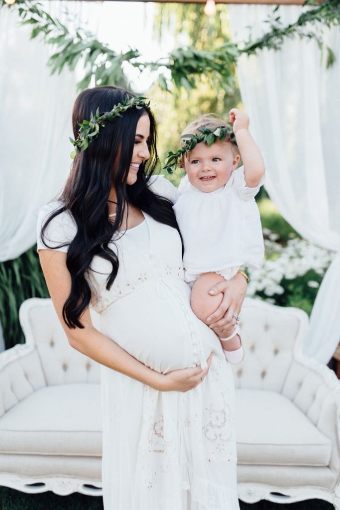 a pregnant woman holding her baby girl in front of a white couch and greenery