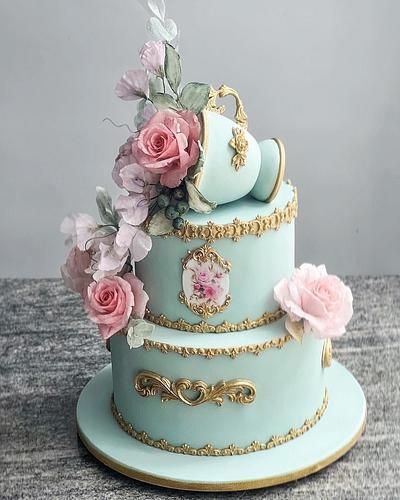 a three tiered blue cake with pink flowers on top and gold trimmings