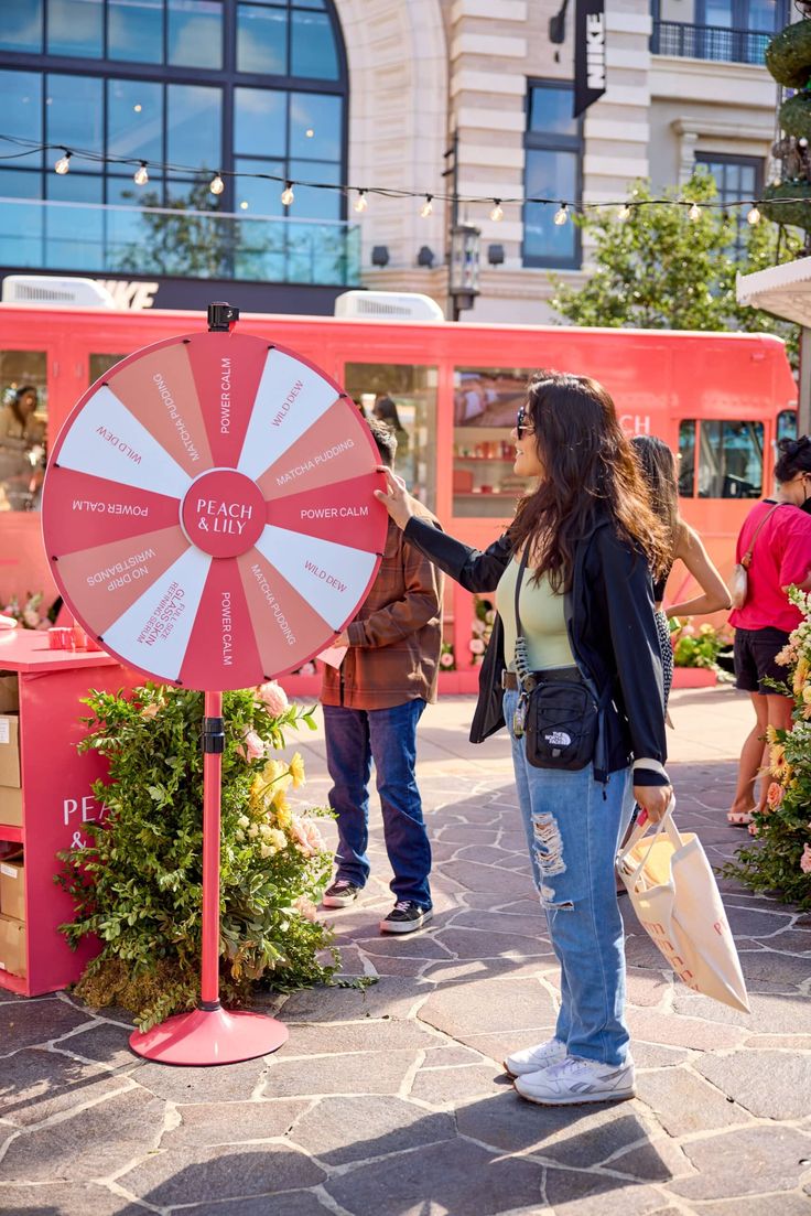 a woman standing in front of a red and white wheel of fortune sign with people looking at it