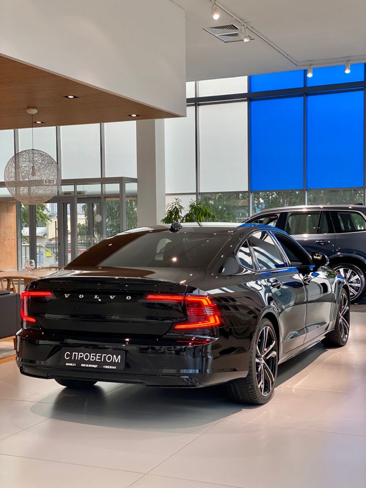 two cars are parked in a showroom with large windows and blue sky behind them