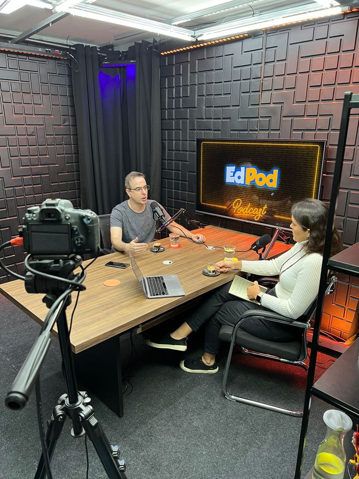 two people sitting at a table with microphones in front of them, talking to each other