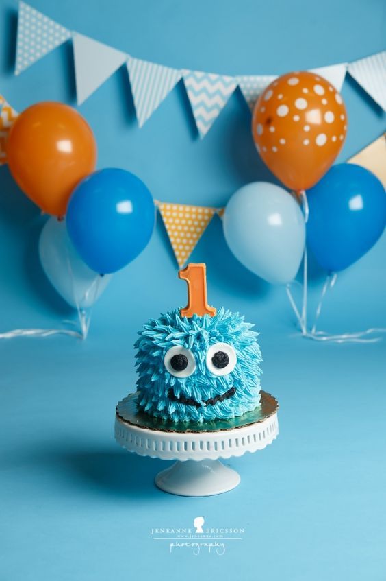 a blue cake with an one on top and balloons in the background for a 1st birthday party