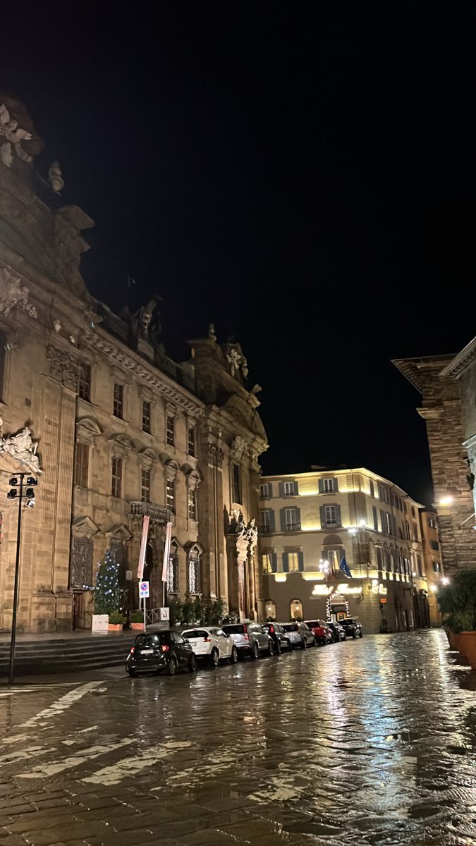 cars are parked on the street in front of an old building at night with its lights on