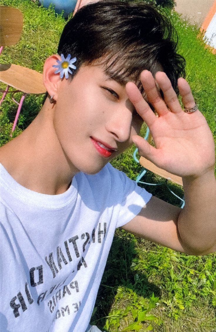 a young man sitting in the grass holding his hand up to his face and wearing a flower in his hair