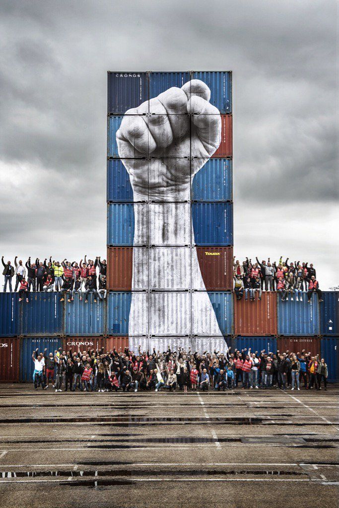 a large group of people standing in front of a tall building with a giant hand painted on it's side