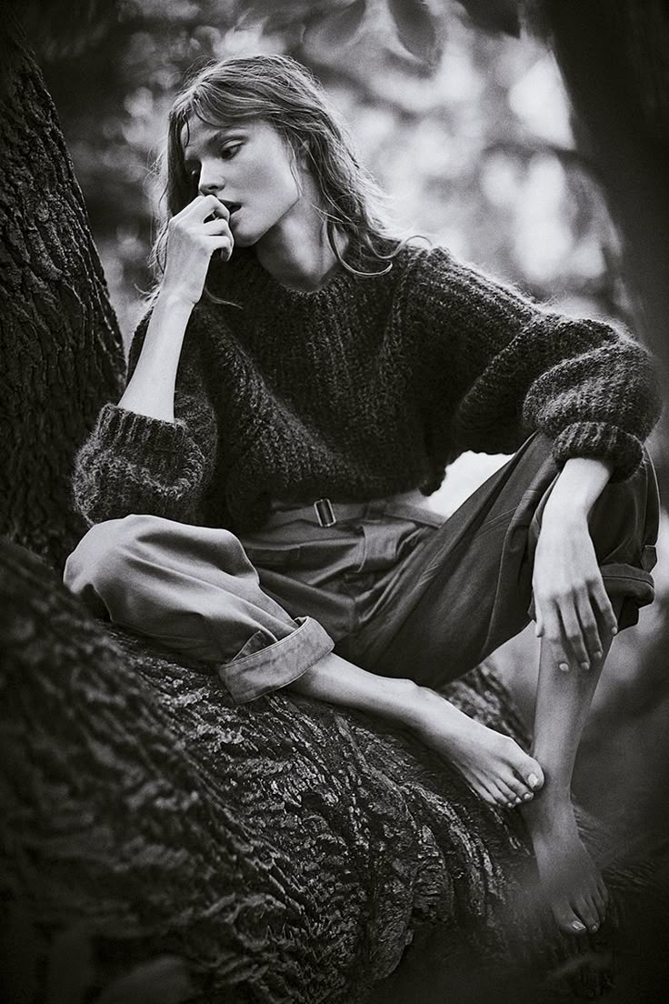 a woman sitting on the ground next to a tree talking on a cell phone with her hand in her mouth