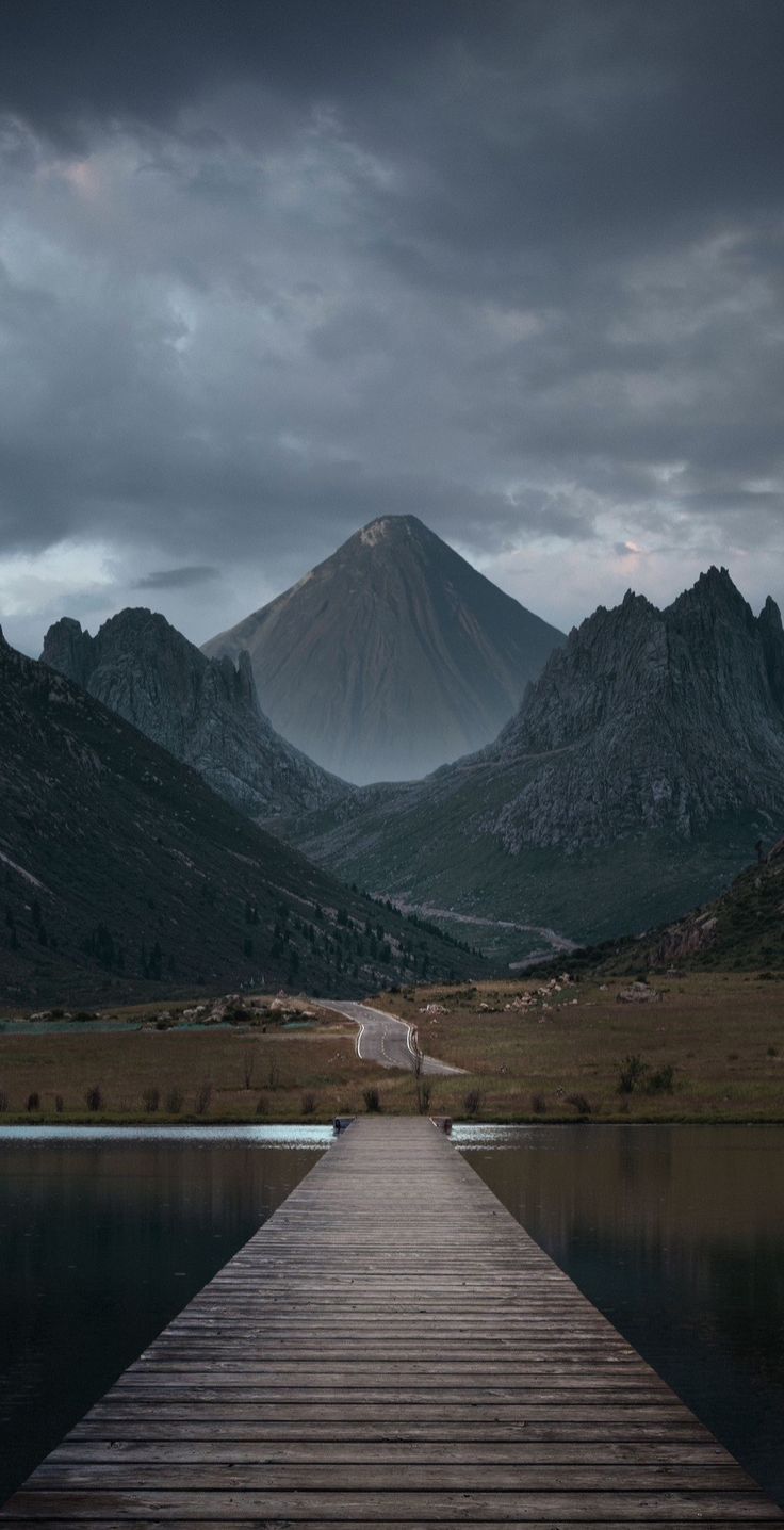 a wooden dock sitting on top of a lake under a cloudy sky with mountains in the background