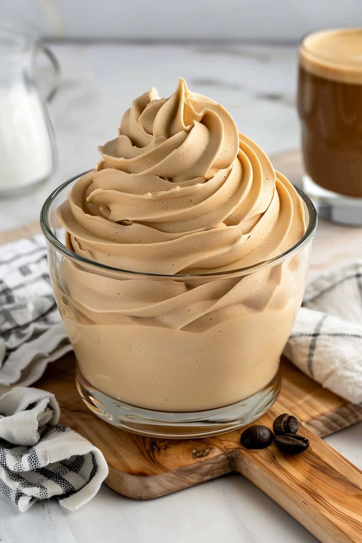 a glass bowl filled with whipped cream on top of a wooden cutting board next to a cup of coffee