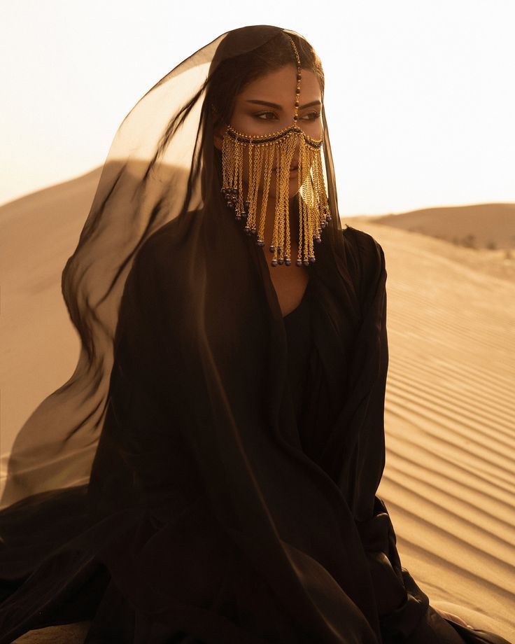 a woman in black dress sitting on top of a sand dune wearing gold jewelry and headdress