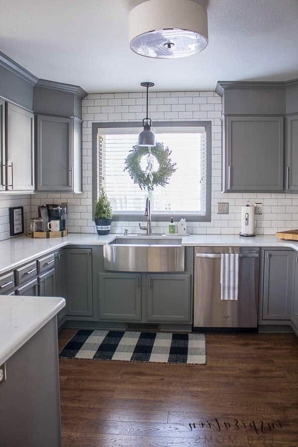 a kitchen with gray cabinets and white tile backsplash, wood flooring and a wreath on the window sill