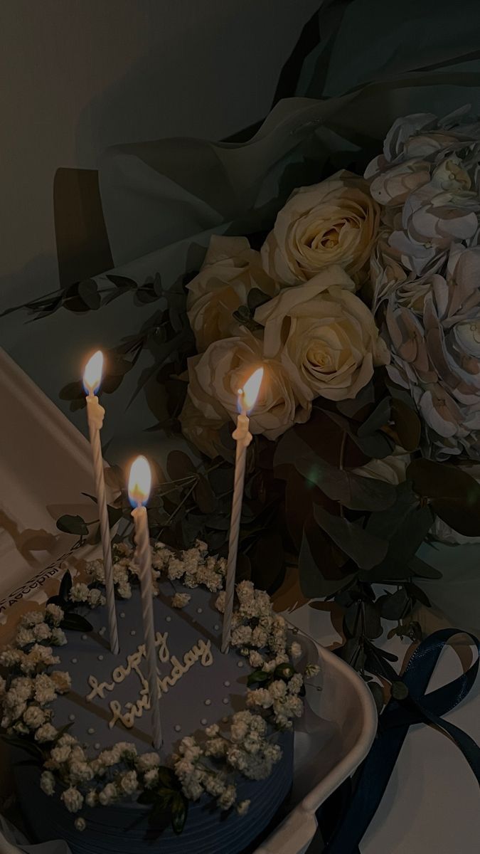 a birthday cake with three lit candles on it and flowers in the backround