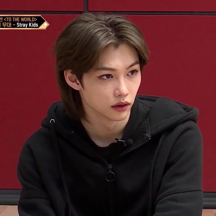 a young man sitting at a table in front of a red wall wearing a black hoodie