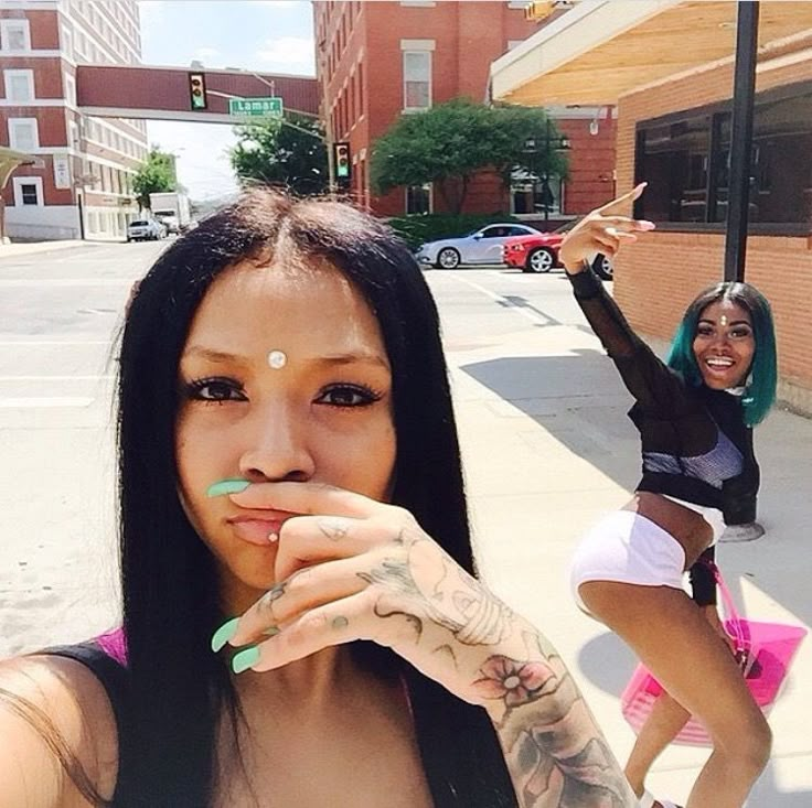 two beautiful young women standing next to each other on a street with one pointing at the camera