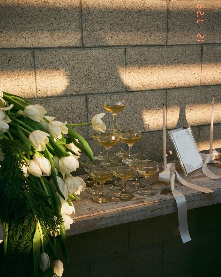 white tulips and wine glasses are sitting on a table next to a brick wall