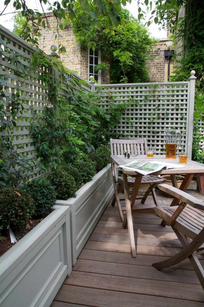 there is a table and chairs on the deck in this backyard area, with plants growing along the fence