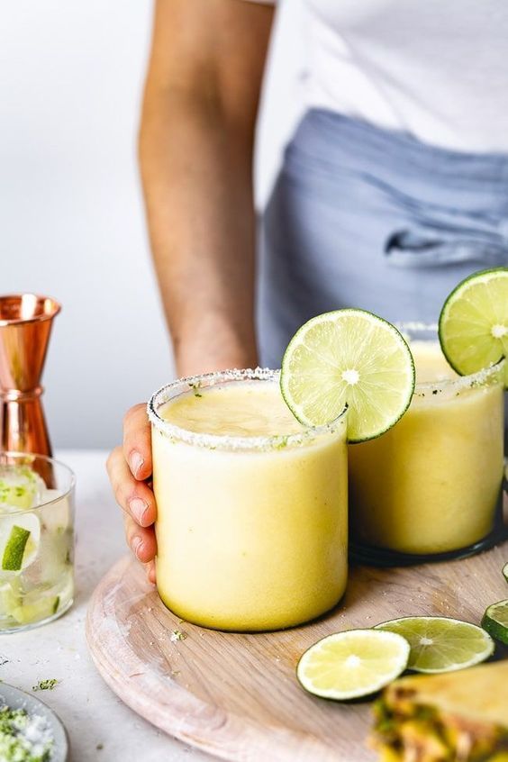 two margaritas with lime wedges on a wooden tray next to copper shakers