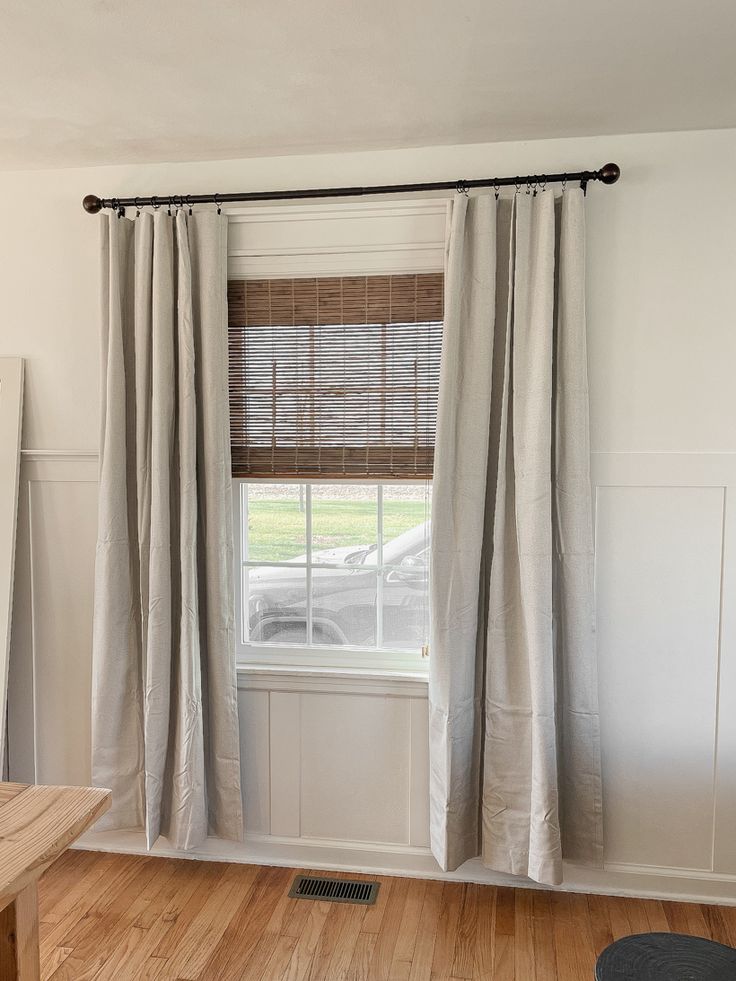 a living room with white walls and wood flooring on the wooden floors, two windows covered in beige drapes