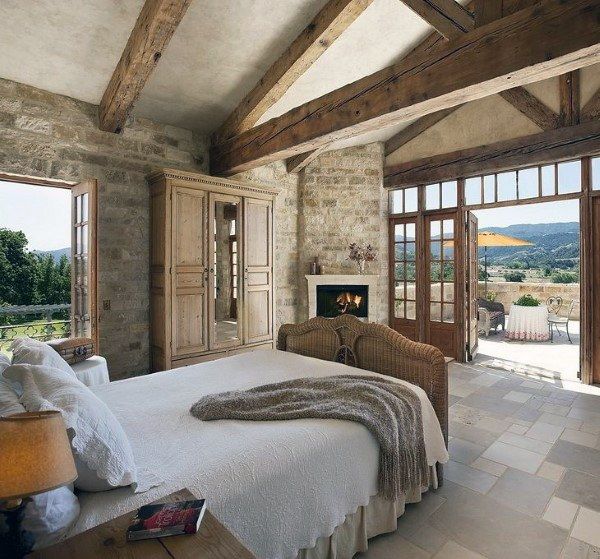 a large bedroom with stone walls and exposed beams, an open fireplace in the center