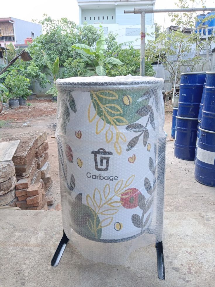 a large trash can sitting on top of a cement floor next to blue barrels and trees