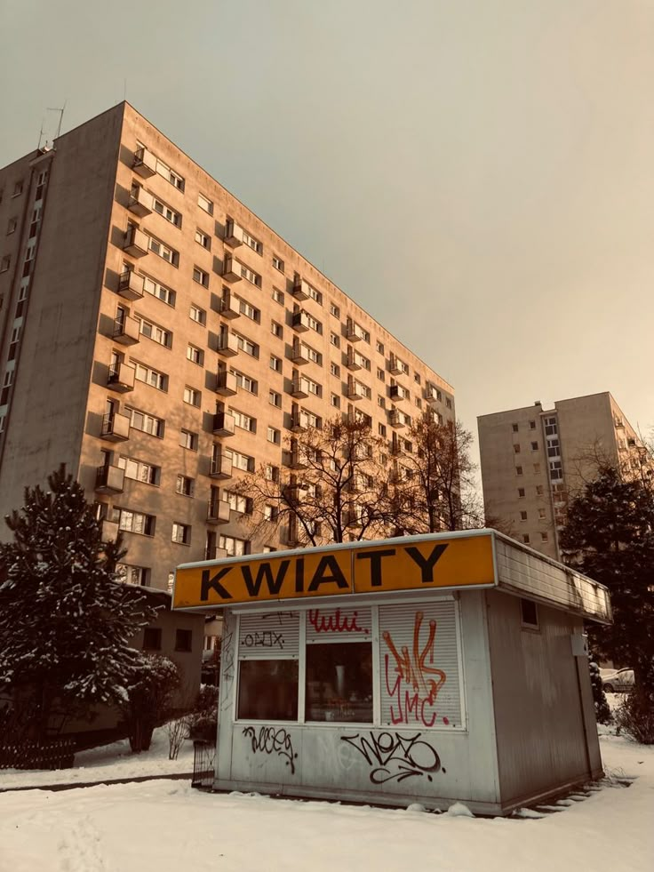 a small building with graffiti on it in the middle of a snow covered area next to tall buildings