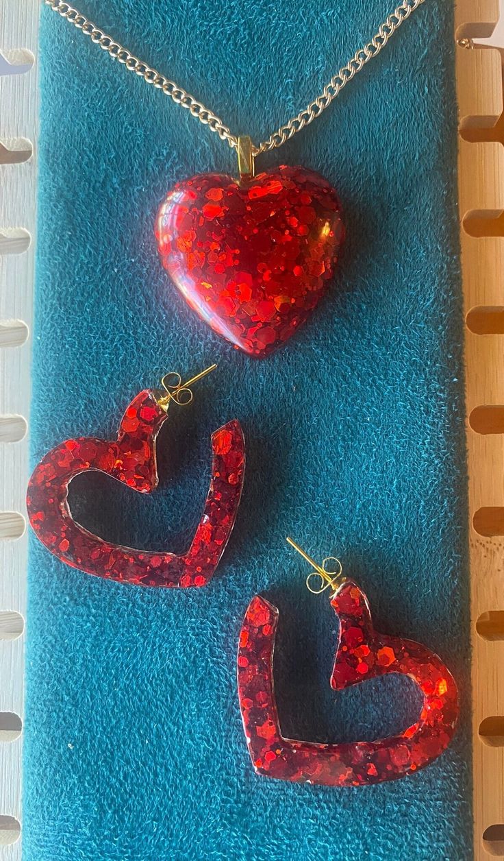 Standout with this vibrant red heart pendant and open heart post back earrings. 18" gold nickle free iron curb chain, 2mm. Lobster clasp closure. Earrings have gold hypoallergenci posts. Made with epoxy resin and red chunk glitter. Handcrafted items often have slight imperfections which are part of the creative process and add to the character of the piece; no 2 are alike!  Colors you see on the screen may differ slightly from in person due to variations in lighting, camera and electronic postin Red Heart Pendant Earrings, Red Open Heart Jewelry For Valentine's Day, Red Pierced Heart Earrings As Gift, Heart Shaped Resin Jewelry For Party, Trendy Resin Jewelry For Valentine's Day, Red Heart Pendant Earrings For Valentine's Day, Valentine's Day Resin Jewelry With Heart Charm, Red Heart Charm Earrings For Party, Red Double Heart Pierced Jewelry