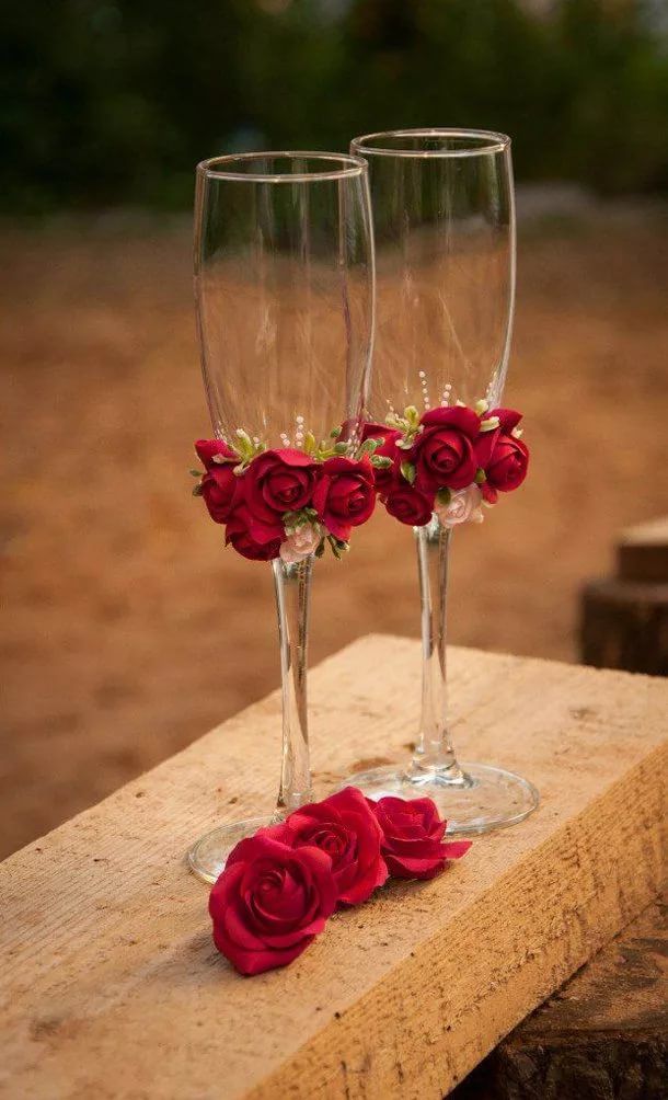 two wine glasses with red roses in them sitting on a piece of wood next to each other