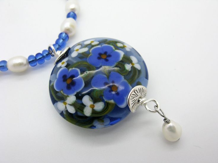 A long necklace with a large handmade flower bead. It is made of a transparent blue glass and decorated decorated with white and blue flowers. Under the large lentil pearl is a movable freshwater cultured pearl. The necklace is threaded with blue transparent glass rocailles  and freshwater cultured pearls. It is closed with a lobster clasp. The circulating length is about 90 cm. In addition, the pendant unit, this measures about 6.5 cm. This necklace goes very well with plain clothes.  Single pi Blue Flower Shaped Beaded Necklace For Gift, Blue Flower-shaped Beaded Necklace Gift, Handmade Blue Flower Pendant Necklace, Plain Clothes, Painting On Glass, Bead Flower, Lampwork Jewelry, White And Blue Flowers, Handmade Lampwork Bead