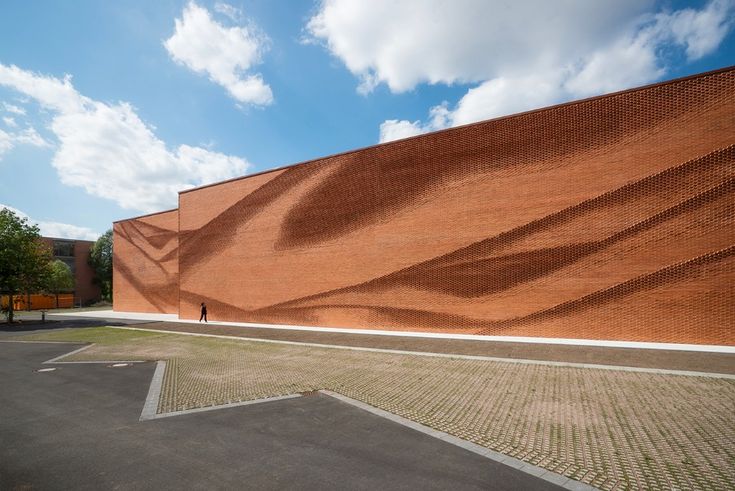 an empty parking lot next to a brick building with shadows on the wall and people walking by