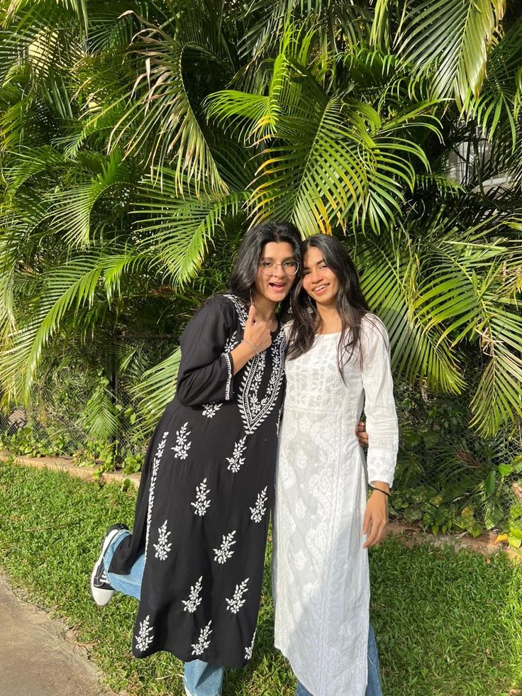 two women standing next to each other in front of some palm trees and grass with their arms around each other